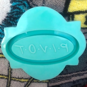 Könnte beinhalten: Eine tealblaue Silikonform in Form eines Banners mit dem Text "T.O.V.I.9" in einem Oval.