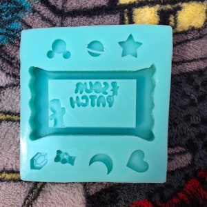 Könnte beinhalten: Eine hellblaue Silikonform mit einem rechteckigen Platz für einen Schokoriegel und mehreren kleineren Plätzen für andere Formen, darunter ein Stern, ein Halbmond, ein Herz, ein Diamant, ein Bonbon und ein Mickey-Maus-Kopf. Der Platz für den Schokoriegel trägt den Text "Sour Patch" in umgekehrter Reihenfolge.