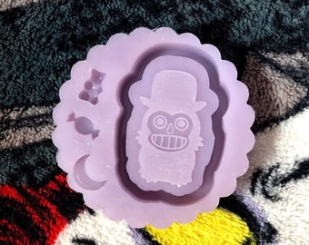 May include: Purple silicone mold with a design of a cartoon ghost wearing a hat and holding a candy cane. The mold also includes a crescent moon and a cluster of three grapes.