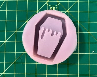 May include: A pink silicone mould with a coffin-shaped design. The coffin has a dripping effect and is outlined in white.