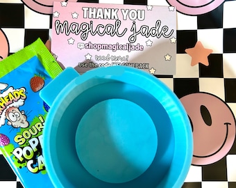 Op de afbeelding: Een blauwe keramische mok met een roze en witte smiley-gezichtsachtergrond. De mok staat op een zwart-wit geblokt oppervlak. Een klein pakje zure snoepjes bevindt zich op de voorgrond. Een bedankkaart met de tekst "THANK YOU magical Jade" bevindt zich op de achtergrond.