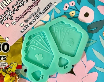May include: A green silicone mold for making resin crafts. The mold is shaped like four playing cards, all aces of clubs, and a single spade. The mold is on a blue and pink floral background.