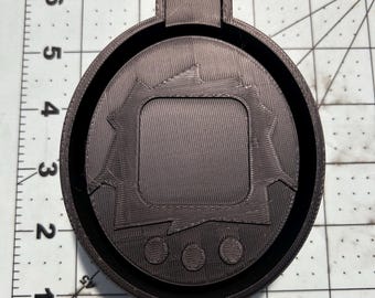 May include: A black plastic mold of a digital pet toy. The mold has a screen, three buttons, and a raised design around the screen.