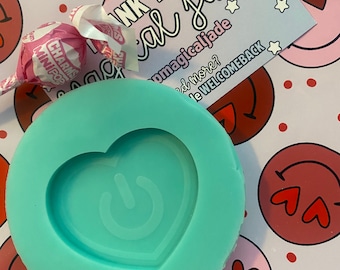 May include: A teal blue silicone mold shaped like a heart with a power button design inside. The mold is on a white background with pink and red hearts and smiley faces.