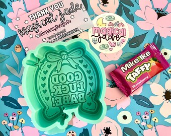 May include: A green silicone mold shaped like a horseshoe with the text "Good Luck Babe!" inside. The mold is on a blue and pink floral background with a pink and white thank you card, a round sticker with the text "magical jade", and a pink and red candy wrapper with the text "Mike Ike Taffy Strawberry".
