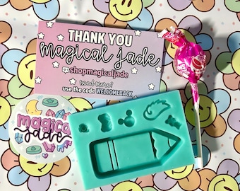 May include: A teal silicone mold with a pencil, a star, a bone, a circle, and a heart design. The mold is on a white background with a pink and blue flower pattern. The text "THANK YOU magical Jade" is on a pink and blue card with white stars.
