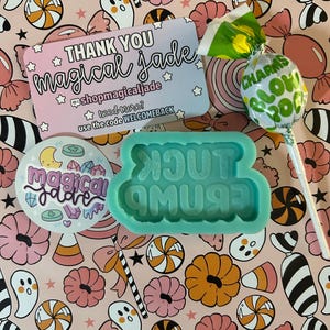 May include: A teal blue silicone mold with the words "You're a**m**u**s**i**n**g" in white letters. The mold is on a pink and white background with a candy theme. There is a business card with the text "THANK YOU magical Jade shopmagicaljade need more? use the code WELCOMEBACK" and a green lollipop with the text "CHARMS BLOW POP" on the background.