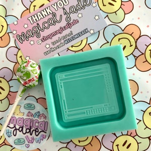 May include: A teal blue silicone mold shaped like a computer screen. The mold is on a white background with a pink, yellow, and blue flower pattern. The text "THANK YOU magical jade" is on a pink and white card with stars and the text "@shopmagicaljade" and "use the code WELCOMEBACK".