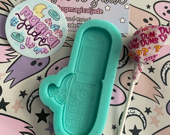 May include: A teal blue silicone mold shaped like a flip phone. The mold is on a white background with a pink, purple, and blue pattern of stars and moons. The mold is for making resin crafts.