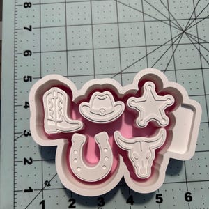 Puede incluir: Un cortador de galletas de plástico blanco con cinco diseños de temática western: una bota de vaquero, un sombrero de vaquero, una estrella de sheriff, una herradura y un cráneo de toro sobre una base de corte con marcas en centímetros.