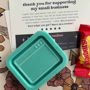 May include: A teal blue silicone mold shaped like a computer screen. The mold is on a white surface with a thank you note that reads "thank you for supporting my small business" and a red candy wrapper that reads "Starburst Fruit Chews".