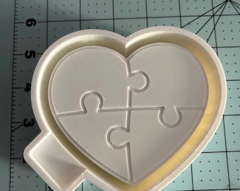 May include: A white 3D-printed heart-shaped puzzle mold. The mold features a heart-shaped outline with a puzzle piece design inside. The puzzle is divided into four pieces. The mold is on a green and white grid surface.