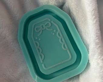 Puede incluir: Un molde de silicona azul claro para manualidades de resina. El molde tiene forma de etiqueta con un lazo y bordes festoneados.