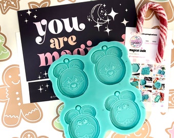 Può includere: Uno stampo in silicone blu turchese con quattro cavità a forma di volti di animali dei cartoni animati. Lo stampo è su uno sfondo bianco con un motivo a biscotti di pan di zenzero. Il testo "you are magic" è visibile su una carta nera dietro lo stampo.