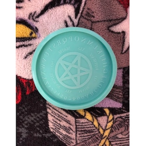 May include: A round, light teal-colored planchette with a raised rim. The planchette features an engraved alphabet, numbers, and the words "yes," "no," and "maybe." A pentagram is in the center. The planchette is a tool for communicating with spirits.