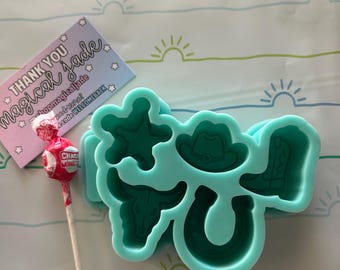 May include: A light turquoise silicone mold featuring western-themed shapes including a sheriff star, a cowboy hat, a bull skull, a horseshoe, and a cowboy boot. A small red and white lollipop and a thank you card with the text Magical Jade are included.