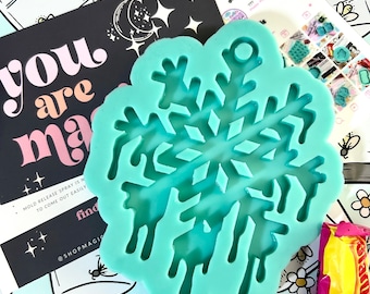 Op de afbeelding: Een turquoise blauwe siliconen mal in de vorm van een sneeuwvlok met een druppel effect. De mal is op een blauwe en witte achtergrond met bloemen en potten. De tekst "you are magic" is zichtbaar op de achtergrond.