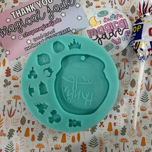 Puede incluir: Un molde de silicona azul turquesa con forma de caldero y otras formas pequeñas, incluyendo una corona, un hongo, una botella, un castillo y una cabeza de Mickey Mouse. El molde está sobre un fondo blanco con un patrón de hongos marrones, hojas verdes y flores naranjas.