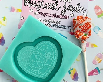 May include: A teal silicone mold shaped like a heart with a design of a cartoon face with hearts for eyes and a telephone inside a heart. The mold is on a blue background with a white and pink pattern. A red and orange lollipop with the words "Orange Dum-Dums" is on the right side of the image.