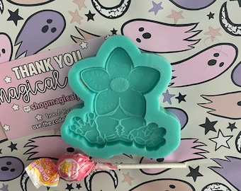 May include: A teal blue silicone mold shaped like a flower with a face. The mold is on a white background with a pink and purple ghost pattern.