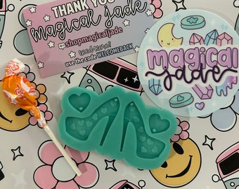 Può includere: Uno stampo in silicone verde a forma di paio di tacchi alti con dettagli a forma di cuore bianco. Lo stampo è su uno sfondo bianco con un motivo floreale rosa e bianco. Il testo "THANK YOU magical jade @shopmagicaljade need more? use the code WELCOMEBACK" è su una carta rosa e blu. È anche visibile un adesivo con il testo "magical jade".