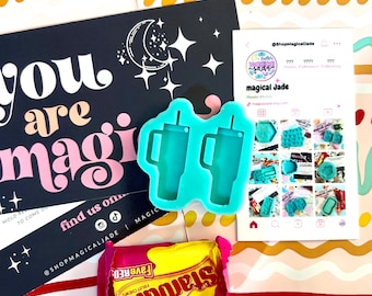 May include: A teal blue silicone mold with two cup shapes. The mold is on a white surface with a pink and yellow background. The text "magical Jade" is visible in the background.