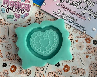 May include: A teal blue silicone mold shaped like a heart with a smaller heart inside. The mold has a beaded border around the outer edge. The mold is sitting on a white background with a pattern of orange and black tigers.