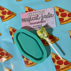 Op de afbeelding: Een teal ovaal hars dienblad met een lolly op een blauwe achtergrond met een herhalend patroon van pizzapunten. De achtergrond bevat ook een roze bedankkaart met de tekst "THANK YOU magical Jade @shopmagicaljade need more? use the code WELCEBACK".