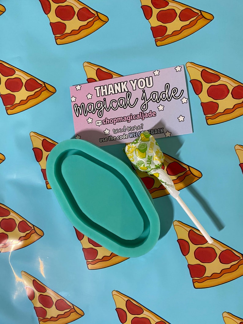Op de afbeelding: Een teal ovaal hars dienblad met een lolly op een blauwe achtergrond met een herhalend patroon van pizzapunten. De achtergrond bevat ook een roze bedankkaart met de tekst "THANK YOU magical Jade @shopmagicaljade need more? use the code WELCEBACK".