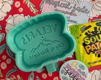 May include: A teal blue silicone mold shaped like a butterfly with the words "Girl of Starlight City" and "VELARIS" written on it. The mold is sitting on a red floral background with a sticker that says "magical jade" and a thank you card that says "Thank you magical jade @shopmagicaljade #comeback".