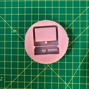May include: A pink silicone mold shaped like a retro computer with a heart on the front. The mold is on a green cutting mat.