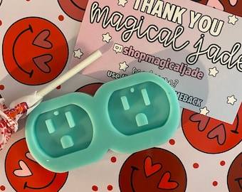 Può includere: Uno stampo in silicone blu chiaro con due forme di prese elettriche. Lo stampo è su uno sfondo bianco con facce sorridenti rosse e rosa con cuori.