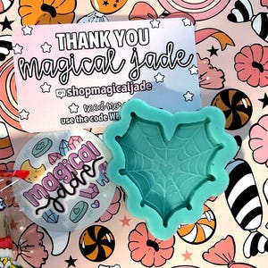 May include: A teal silicone mold in the shape of a heart with a spiderweb design, a round sticker, and a thank you card with the text "magical jade" on a colorful candy-themed background.