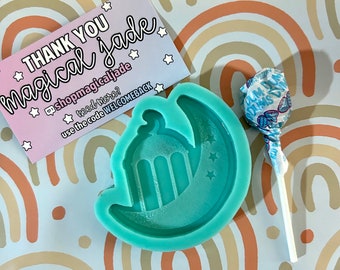 May include: A teal blue silicone mold shaped like a crescent moon with a star and a mosque inside. The mold is on a white background with rainbow arches.