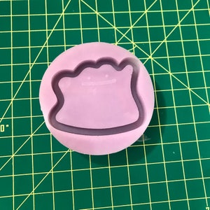 May include: A pink silicone mold with a raised outline of a cartoon character. The character has a round body, two small arms, and a large head with a wide smile.