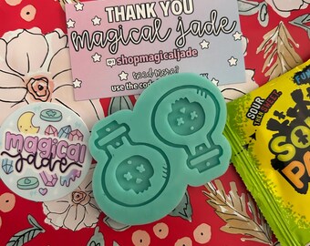 Könnte beinhalten: Eine tealblaue Silikonform mit zwei Formen für Zaubertrankflaschen. Die Form befindet sich auf einem roten Hintergrund mit weißen Blumenmustern. Der Text "THANK YOU magical jade" befindet sich auf einer rosa und blauen Karte mit weißen Sternen.