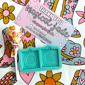 May include: A turquoise silicone mold with two square compartments, each with a circular cutout. The mold is for making resin earrings. The mold is on a white background with a pink and blue cowboy boot pattern, a pink cowboy hat with smiley faces, and a lollipop.