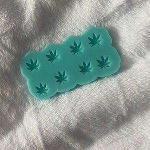 May include: A light blue silicone mold with a design of multiple marijuana leaves. The mold is on a white fluffy surface.