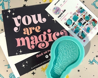 May include: A teal silicone mold in the shape of a lightbulb with a floral design. A black card with the words "you are magic" in pink and peach. A business card with social media information.