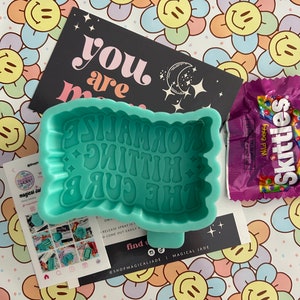 Puede incluir: Un molde de silicona azul turquesa con forma de cartel con el texto "The Curb Hitting Normalise" en una fuente retro. El molde está sobre un fondo blanco con un patrón de flores de colores.