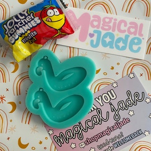May include: A teal blue silicone mold shaped like two inflatable flamingo pool floats. The mold is on a white background with a rainbow pattern. A pink and blue sticker with the text "Magical Jade" is in the background. A thank you card with the text "Thank you magical jade" is in the foreground. The card has a pink background with white stars and the text "@shopmagicaljade" and "use the code WELCOMEBACK".