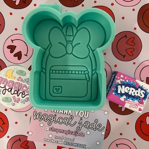 May include: A green silicone mold shaped like a backpack with a bow on top. The mold is on a pink and white background with hearts and smiley faces. The text "THANK YOU magical Jade" is on a blue and white card with stars. The text "@shopmagicaljade" is on the card. The text "need more? use the code WELCOMEBACK" is on the card.