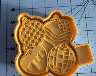 May include: A yellow plastic cookie cutter featuring four Christmas ornament designs. The ornaments include a checkered pattern, wavy lines, a bow, and a heart with dripping details. The cutter has a handle for easy use.