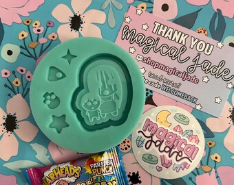 May include: A silicone mold with a design of two cartoon characters, a star, and a crescent moon. The mold is on a blue and pink floral background with a pink and white candy wrapper and a sticker that says "magical jade".
