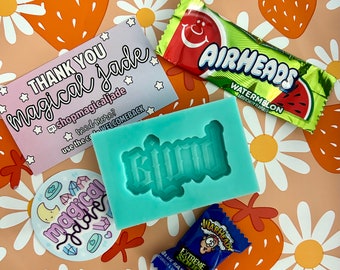 May include: A small business thank you card with the text "Thank You magical jade" and a star design. The card is on a pink and white background with a daisy and strawberry pattern. There is a blue silicone mold with the word "Girl" in it. There are two small candy wrappers, one blue and one green.