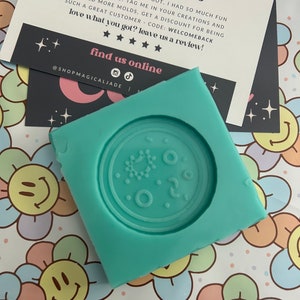 Puede incluir: Un molde de silicona azul turquesa con forma de círculo con un diseño de círculo más pequeño en el interior. El molde está sobre un fondo blanco con flores de colores.