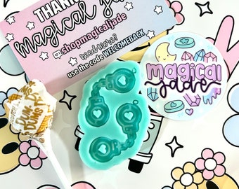 Può includere: Un biglietto di ringraziamento con il testo "THANK YOU magical jade" e il nome del negozio "shopmagicaljade". Il biglietto è su uno sfondo bianco con fiori rosa e bianchi, stelle e un viso sorridente. C'è anche uno stampo in silicone blu di manette con cuori sulle catene, un lecca-lecca e un adesivo con il testo "magical jade" e una luna, stelle e cuori.