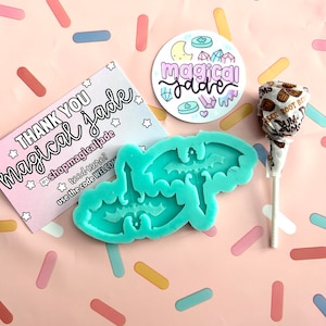 May include: A teal blue silicone mold shaped like two bats with a crescent moon in the center. The mold is on a pink and white background with sprinkles. A sticker with the text "magical jade" is in the background.