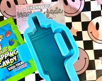 May include: A light blue silicone mold in the shape of a bottle. The mold is on a black and white checkered background with pink smiley faces and star shapes. A packet of candy and a card that says "THANK YOU magical jade" are also visible.