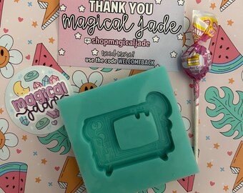 May include: A teal silicone mold in the shape of a retro television, a lollipop, and a sticker with the text "magical jade" are displayed on a pink patterned background. A business card with the text "THANK YOU magical jade" is also visible.